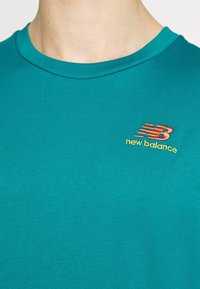 Tyrkysové tričko s krátkým rukávem vyrobené z bavlny, s kulatým výstřihem a vyšitým logem New Balance v oranžové a žluté barvě.
