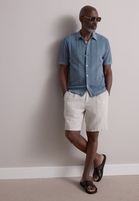 Jasnoniebieska koszula z krótkim rękawem z teksturowanym wzorem diamentowym, beżowe szorty i brązowe sandały z krzyżującymi się paskami. Okulary przeciwsłoneczne dopełniają stylizację.