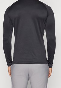 Musta pitkähihaisella urheilupaidalla, jossa on sileä rakenne, litteät saumat ja tiukka muotoilu, yhdistettynä vaaleanharmaisiin urheiluhousuihin.