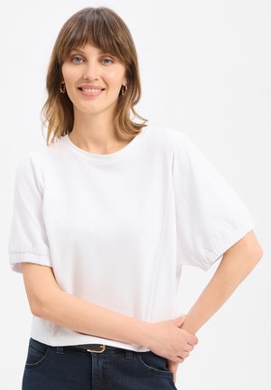 Frau mit schulterlangem braunem Haar, die goldene Creolen, eine weiße kurzärmelige Bluse und dunkle Jeans trägt, lächelnd mit einer Hand auf der Hüfte.