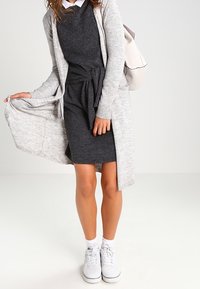 Abito grigio a maglia con vita legata, indossato sotto un cardigan grigio chiaro. Abbinato a scarpe da ginnastica bianche e calzini alla caviglia. Design semplice e casual.