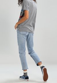Persona vestida con una camiseta gris, jeans cortos de mezclilla claros, calcetines a rayas y zapatillas deportivas azul marino, avanzando sobre un fondo simple.