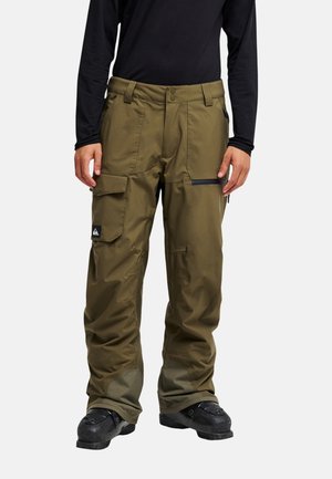 Man draagt olijfgroene cargobroek met zwarte laarzen en een zwart shirt met lange mouwen, staat met ontspannen handen langs het lichaam.
