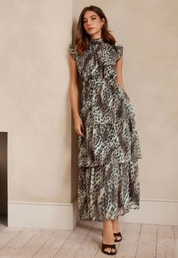 Tiered maxi dress in green and brown animal print. Features ruffled sleeves and a high neckline. Soft fabric, relaxed fit. Worn with brown heels.