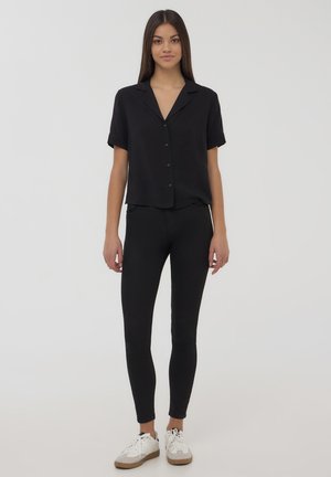 Camicia nera a maniche corte con bottoni abbinata a leggings neri. Il modello indossa scarpe da ginnastica bianche con dettagli beige. Tessuto morbido, vestibilità sartoriale.