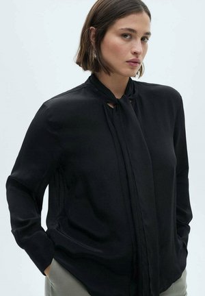 Black long-sleeve blouse in smooth fabric, featuring a tie detail at the collar and side pockets. Subtle sheen and relaxed fit.