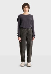 Suéter gris oscuro, acanalado, con escote amplio, combinado con unos jeans negros de alta cintura y corte holgado. La modelo lleva zapatillas beiges.