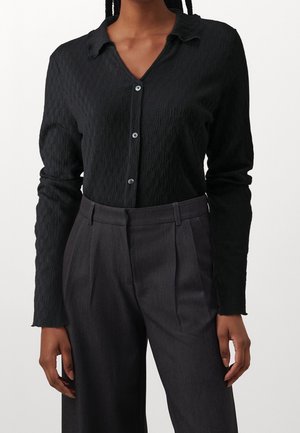 Femme portant une chemise noire texturée à manches longues boutonnée, rentrée dans un pantalon plissé gris foncé taille haute.