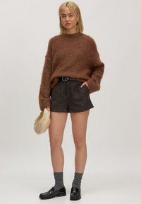 Frau mit blonden Haaren, die einen braunen, wuscheligen Pullover, einen schwarzen Gürtel, dunkle strukturierte Shorts, graue Socken und schwarze Slipper trägt und eine beige Clutch-Tasche hält.
