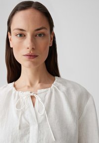 Blusa bianca in lino con scollatura rotonda, dettagli arricciati e chiusura a legatura. Il tessuto ha una finitura morbida e testurizzata.