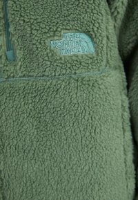 Zeleni polar pulover s mekanom, teksturiranom površinom. Ima ovratnik s polu-zipom i utisnutim logotipom svijetloplave boje na prsima.