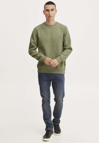 Felpa verde oliva con scollatura a girocollo, polsini e orlo a costine. Indossata con jeans blu e sneakers nere. Design minimalista con dettaglio del logo.