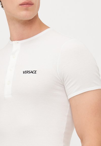 Maglietta henley bianca a maniche corte con costine, realizzata in un materiale elastico, con un'apertura a bottoni e un piccolo logo nero "VERSACE" sul petto.