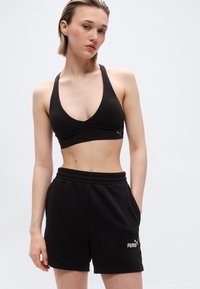 Γυναίκα με μαύρο αθλητικό σουτιέν και μαύρο σορτς Puma, που στέκεται με το ένα χέρι στην τσέπη, μπροστά σε απλό φόντο.