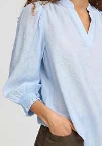 Blouse bleu clair à col, avec une finition texturée. Présente des manches élastiques froncées. Associée à un pantalon vert olive.