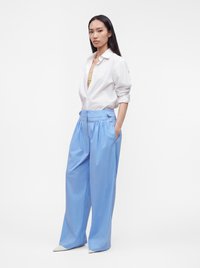 Femme aux longs cheveux foncés portant une chemise blanche boutonnée et un pantalon large bleu clair taille haute, debout les mains dans les poches.