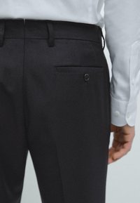 Zwarte broek met een soepele textuur heeft een enkele achterzak met een drukknopsluiting. Een witte overhemden is gedeeltelijk zichtbaar.
