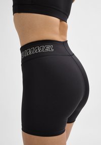 Zwarte sportshorts van rekbaar materiaal, met een hoge tailleband waarop het woord "HUMMEL" in witte letters langs de bovenkant staat.