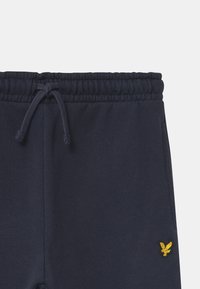 Marina blå träningbyxor med resårmidja och snörning samt en liten guldkopparörn-logotyp på vänster ben. Mjuk material, minimalistisk design.