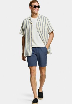 Man met zwarte zonnebril, een kort mouw gestreept overhemd over een wit T-shirt, blauwe shorts en zwarte instapschoenen, lopend met een glimlach.
