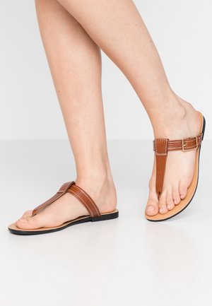 Pieds portant des sandales marron en cuir à brides en T avec fermetures à boucle, présentés sur un fond clair uni.
