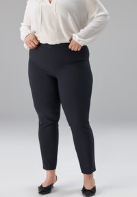 Schwarze, schmal zulaufende Hose aus glattem Stoff, kombiniert mit einer weißen Bluse mit V-Ausschnitt und gerafften Manschetten.