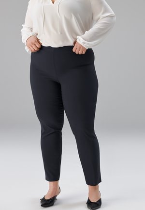 Schwarze, schmal zulaufende Hose aus glattem Stoff, kombiniert mit einer weißen Bluse mit V-Ausschnitt und gerafften Manschetten.