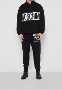 Černá mikina Moschino s bílým logem na hrudi, doplněná černými tepláky se postranním logem. Oba kusy mají volný střih.