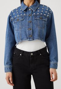 Lyhyt sininen farkkutakki, jossa on hopeiset niitti koristeet harteilla, edessä napitus ja kaksi rintataskua. Päällä valkoinen paita.