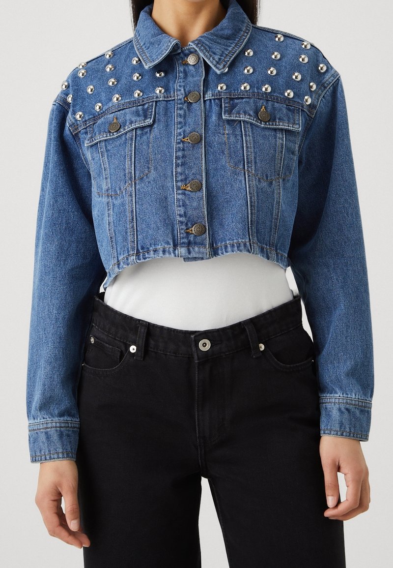 Lyhyt sininen farkkutakki, jossa on hopeiset niitti koristeet harteilla, edessä napitus ja kaksi rintataskua. Päällä valkoinen paita.