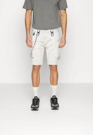 Pantaloni corti beige chiaro con tasche cargo, caratterizzati da dettagli neri, cinghie regolabili e una trama liscia. Abbinati a una maglietta grigia e scarpe da ginnastica.
