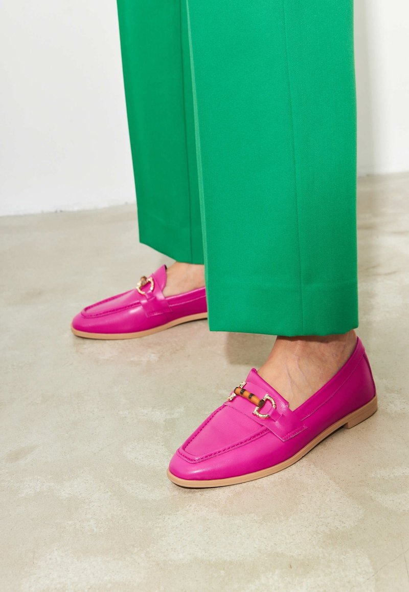 Pieds portant des mocassins en cuir rose vif avec des détails dorés et en bambou, assortis à un pantalon vert à jambes larges sur un sol neutre.