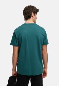 Teal bomull T-skjorte med korte ermer, rund hals, og en liten merkelapp på baksiden, holdt i hånden ved siden av en mørk caps.