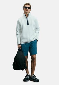 Veste zippée blanche avec un col haut, associée à un short bleu et des baskets noires. Tenant un sac noir, se tenant devant un fond blanc.