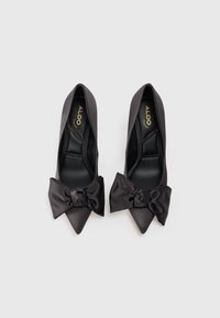 Scarpe nere in satin con tacco alto e punta affilata, caratterizzate da un grande fiocco decorativo sulla tomaia e una texture liscia. Nome del marchio visibile all'interno.
