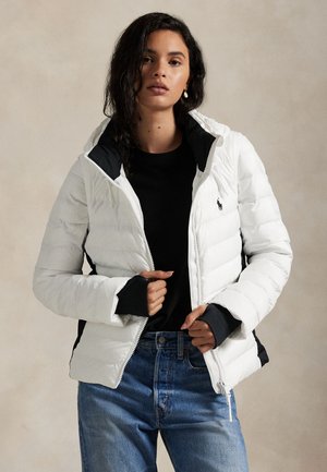 Donna che indossa una giacca imbottita bianca sopra una maglia nera e jeans blu, con le mani vicino alla cerniera della giacca, sfondo neutro.