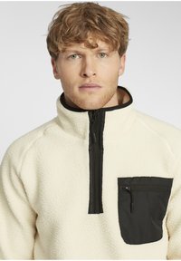 Cream fleece pullover with a high collar, featuring a black zippered chest pocket and black zipper trim, providing a soft, textured finish.