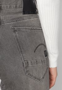 Šedé denimové kalhoty s zakřivenou zadní kapsou a černou koženou cedulkou. Tkanina se zdá být texturovaná s jemnými detaily šití.