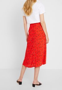 Jupe midi rouge à fleurs en tissu léger avec une taille ajustée, s'évasant vers l'ourlet. Des sandales noires complètent la tenue.