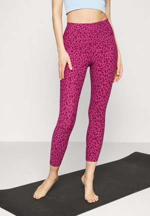 Vrouw staat blootsvoets op een zwarte yogamat, draagt hoog getailleerde roze legging met donkere luipaardprint en een lichtblauwe cropped top.