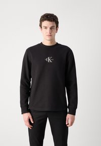 Fekete, hosszú ujjú pulóver bordázott mintázattal, kerek nyakkal és fehér Calvin Klein logóval a középen. Fekete nadrággal párosítva.