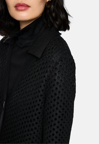 Schwarze, strukturierte Strickjacke mit einem gemusterten Gewebe, ausgestattet mit einem kontrastierenden einfarbigen Kragen und Bündchen, kombiniert mit einem schwarzen Oberteil.