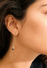 Goudkleurige dangle oorbel met een lange ketting, met parel en zwarte kraal accenten, gedragen aan het oor van een model met donker haar.
