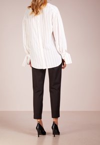 Femme portant une blouse blanche avec de fines rayures rouges, un pantalon noir ajusté et des chaussures à talons noirs, se tenant sur un sol neutre.