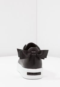 Sneaker in pelle nera con suola bianca testurizzata, caratterizzato da un grande fiocco in satin nero sul tallone e un logo impresso sul retro.