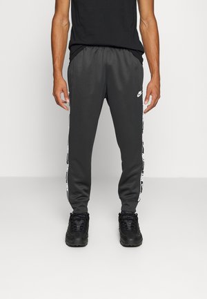 Graue Sporthose aus glattem Stoff, mit weißen Seitenstreifen und Logo-Akzenten sowie umgeschlagenen Säumen, kombiniert mit schwarzen Sneakers.