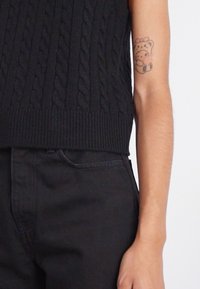 Black cable-knit sleeveless top with a ribbed hem, paired with black denim pants. Notable tattoo of a bear with a bow on the upper arm.