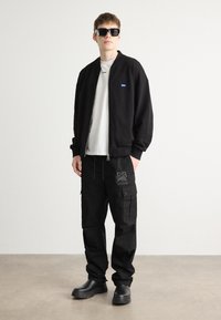 Schwarze Bomberjacke mit blauem Logo, weißes T-Shirt, schwarze Cargo-Hosen mit grauer Markenkennzeichnung und schwarze Fußbekleidung. Sonnenbrillen runden den Look ab.