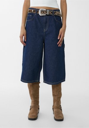 Henkilöllä on yllään tummansiniset polveen ulottuvat denimshortsit, joissa on niitattu leopardikuvioinen vyö, valkoinen yläosa ja polven korkeudelle ulottuvat ruskeat mokkasaappaat.