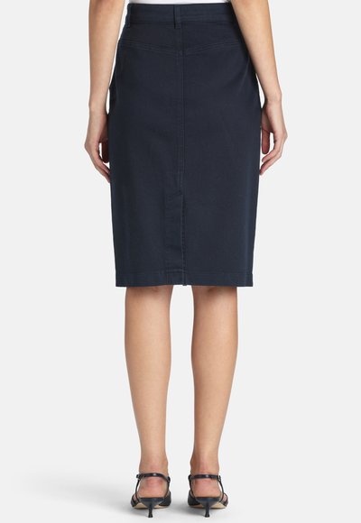 Kvinde iført en knælængde mørk marineblå pencilnederdel og sorte lavhælede sandaler, vist bagfra.